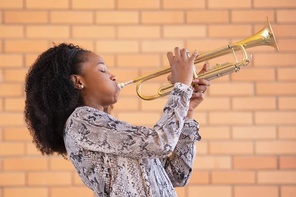 Joven mujer afroamericana tocando la trompeta con los ojos cerrados sobre un fondo de pared de ladrillo Fotos de stock
