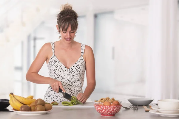 Mulher adulta jovem cortando fatias de kiwi e tendo frutas diferentes ao redor ao café da manhã — Fotografia de Stock