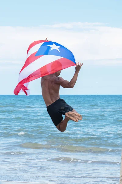 Vista trasera de un hombre sosteniendo una bandera cubana mientras salta a la playa. Fotos de stock libres de derechos