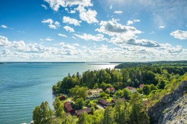 Güzel doğa manzarası. Göl yüzeyinde yeşil ağaçlar ve mavi gökyüzü arka planıyla çevrili binalar. İsveç.