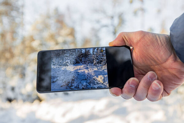 Beautiful view of male hand holding mobile and taking beautiful winter landscape picture. Sweden.