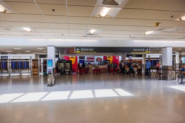 Bright interior view of airport terminal with flight information boards, small cafe and walking passengers. Miami. USA. 