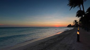 Kartal Sahili sahil şeridinde günbatımı manzarası sakin dalgalar ve palmiye ağaçları tropikal akşam arkaplanı oluşturuyor. Aruba.