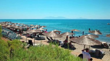 Saman şemsiyeli güneşli çakıl taşı plajı ve adada Ege Denizi kıyısında dinlenen turistler. Kos. Yunanistan. 
