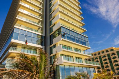 İşçiler, mavi gökyüzü ve güneş ışığının arkasındaki palmiye ağaçlarının yakınındaki modern otel binasının cam cephelerini temizliyorlar. Cancun. Meksika. 