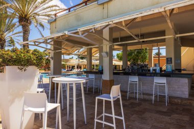 Açık havuz kenarındaki barın beyaz sandalyeli ve arka plandaki tropik tatil beldesinin parlak güneş ışığıyla yakın görüntüsü. 