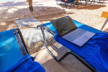 Dizüstü bilgisayarın mavi güneşli yatağını, arka planda plaj bölgesi olan otel avlusunda havluyla kapatın.. 