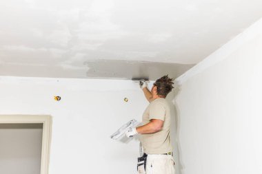 İnşaat işçisinin iç daire tadilatı sırasında geniş spatula kullanarak tavan köşesindeki taze alçıyı düzleştirmesini yakından izleyin..