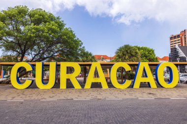 Curacao şehir simgesinin yakın görüntüsü mavi gökyüzüne karşı tropikal ağaçlarla işaretlenmiş sarı harfler..