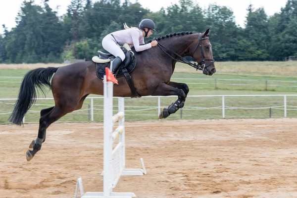 Jovem loira de cabelos compridos cavalo pulando um cavalo escuro — Fotografia de Stock