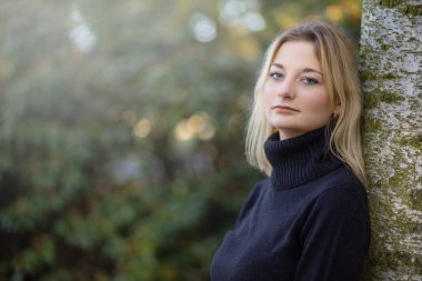 Balıkçı yaka giyen havalı genç kız sonbahar parkında kameraya bakarak poz veriyor. Yatay olarak. 