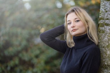 Balıkçı yaka giyen güzel genç kız sonbahar parkında kameraya bakarak poz veriyor. Yatay olarak. 