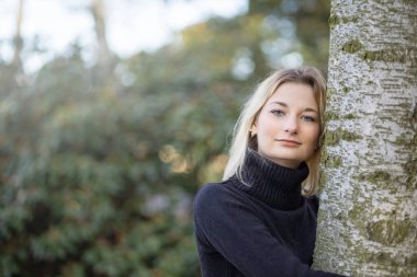 Balıkçı yaka giymiş ciddi bir genç kız sonbahar parkında bir ağaca yaslanmış kameraya bakıyor..