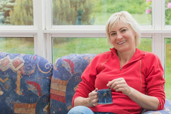 Smiling blonde middle-aged woman is relaxing on a sofa — Stock Photo, Image