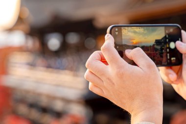 Japonya 'nın Kyoto kentindeki Kiyomizu-dera tapınağındaki akıllı telefondan fotoğraf çeken turistin yakın çekim ve arka planında doğal kırmızı akçaağaç yaprakları ve günbatımı gökyüzü arka planı var..