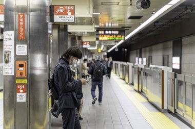 Osaka Şehri, Japonya - Aralık 06 2024: Japon gençlerin metroda bekleyen trenler arasında akıllı telefon kullanışına yakın çekim ve bakış açısı.