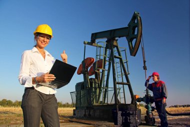 Petrol sahasında çalışan işçiler, elinde not defteriyle gülümseyen kadın mühendis başparmağından vazgeçiyor ve petrol işçisi petrol pompası ve takım çalışmasını kontrol ediyor.