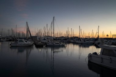 VILANOVA I LA GELTRU, İSPA - 8 ARALIK 2023: günbatımında limana yanaşmış yelkenli ve yatlarla huzurlu bir liman. Durgun su renkli gökyüzünü yansıtırken, direkler mavi ve turuncunun gradyanı karşısında siluetler oluşturur.