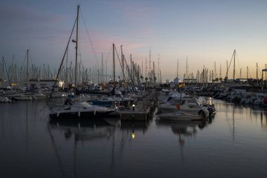 VILANOVA I LA GELTRU, İSPA - 8 ARALIK 2023: günbatımında limana yanaşmış yelkenli ve yatlarla huzurlu bir liman. Durgun su renkli gökyüzünü yansıtırken, direkler mavi ve turuncunun gradyanı karşısında siluetler oluşturur.