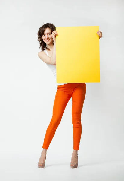 Joyful young woman in orange pants holding blank yellow placard — Stock Photo, Image