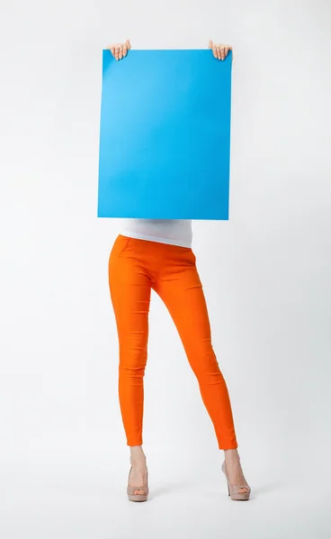 Young woman in orange pants holding blank blue placard hiding b — Stock Photo, Image