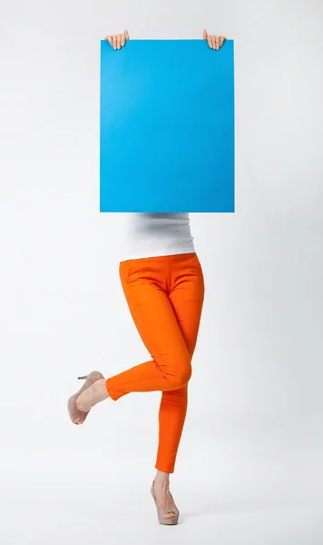 Jovencita juguetona en pantalones naranjas sosteniendo cartel azul en blanco h —  Fotos de Stock