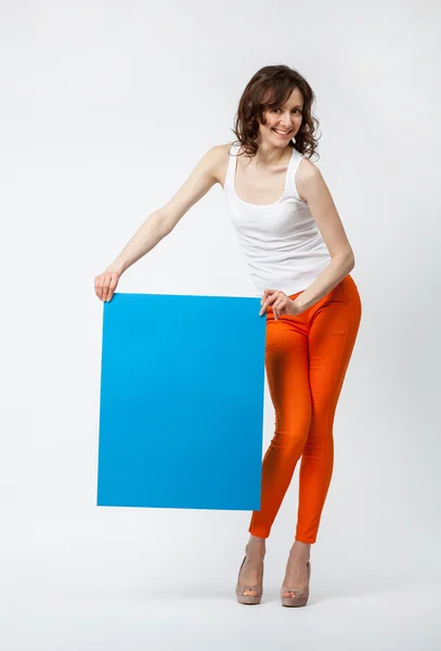 Jovencita juguetona en pantalones naranjas sosteniendo cartel azul en blanco —  Fotos de Stock
