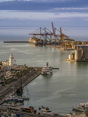 Günbatımında bir Akdeniz limanının panoramik görüntüsünde vinçlerle yüklü büyük bir konteyner gemisi, lüks yatlı marina, beyaz deniz feneri ve liman sanayi tesisleri, Malaga, İspanya