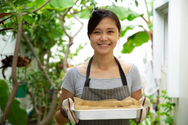 Asyalı temizlikçi kadın elinde bovling tepsisi tutuyor. Ev hanımı aile için yemek yapıyor ve bahçede duruyor..