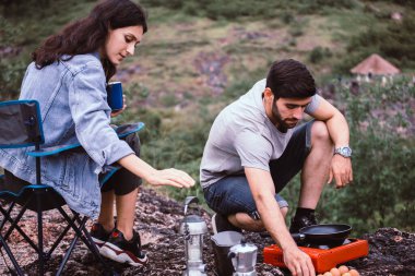 Bir çift aşığı çadırın dışında yemek pişiriyor, kamp kavramının tadını çıkarıyor.