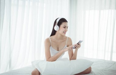 Güzel, mutlu bir kadın cep telefonu ve kulaklık kullanıyor yatak odasında dans ederek müzik dinliyor ve dinleniyor.