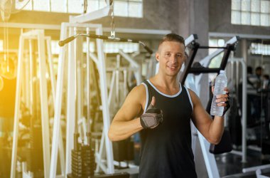 Çekici yakışıklı adam, başparmağını salonda gösterip su içiyor, erkek molası veriyor ve spor yaptıktan sonra dinleniyor.