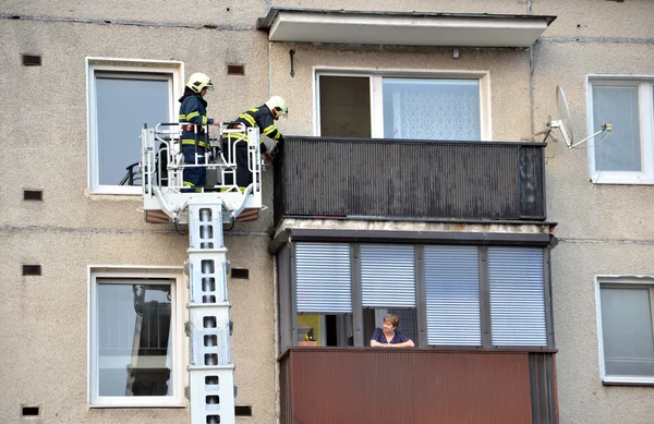 Due pompieri nel cestino telescopico del camion dei pompieri cercano di arrivare al balcone piatto. Vecchia donna li sta guardando dal balcone accanto . — Foto Stock