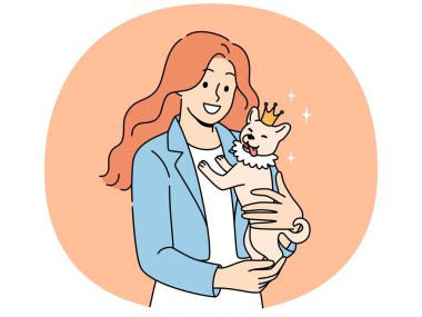 Kadın, kucağında taç takmış küçük bir köpek tutuyor ve gülümsüyor. Yavru köpeklerin koruyuculuğundan keyif alıyor. Mutlu kız hayvanları sever safkan köpeğin huzurunda sevinir ve gülümseyerek ekrana bakar.