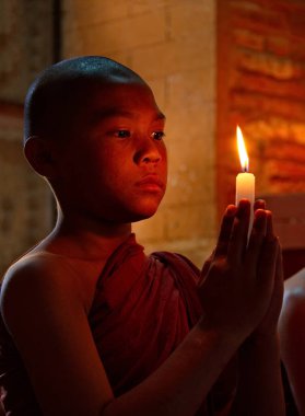 Bagan. Myanmar. 26 Kasım 2016. Ozan rengi kahaya giymiş bir çocuk elinde mumla Budist tapınağında oturan bir Buda heykelinin önünde dua okuyor..