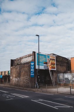 Brentford, Batı Londra (124; İngiltere - 2021.03.21): İşçi yeni reklam panosu için ilan panosu hazırlıyor. Endüstriyel tırmanışçı bir merdiven üzerinde çalışıyor - bir reklam pankartı yerleştiriyor.