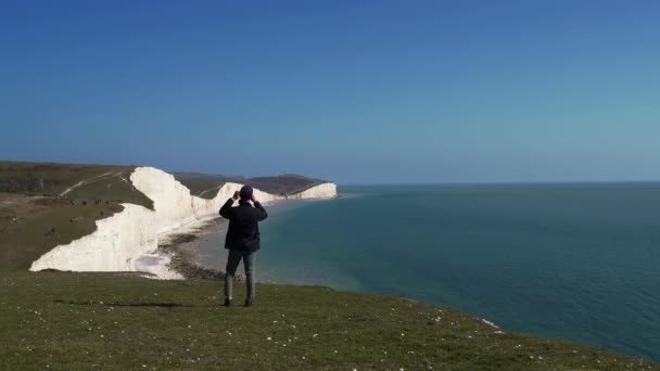 Людина йде до краю мису Семи сестер, щоб сфотографувати Вид на крейдяні скелі і море Ла-Маншу з краю. Південь Англії — стокове відео