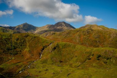 Kardan Donya, Galler 2025.09.20 Çimenli yamaçların ve engebeli kaya oluşumlarının üzerinde yükselen güneşli bir tepe Eryri Ulusal Parkı 'nın açık mavi gökyüzü altında. 