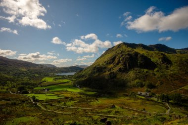 Kardan Donya, Galler 2025.09.21 Parlak yeşil vadi Işıldayan bir göl ve berrak mavi bulutların altında dik dağ yamaçları. 