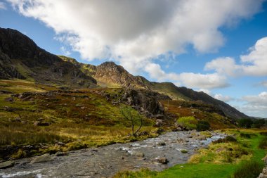 Kardan Donya, Galler 2025.09.21 Berrak bir dağ deresi, engebeli tepeler ve bulutlu mavi gökyüzünün altındaki kayalık tepelerle çevrili yeşil bir vadi boyunca esiyor.. 