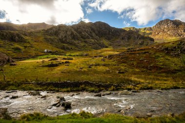 Snowdonia, Galler 2025.09.21 Engebeli tepelerin arasında huzurlu bir taş kulübe ve dramatik bulutların altında akan bir dağ deresi ve uzak bir Galler vadisinde güneş ışığı bulunur.. 