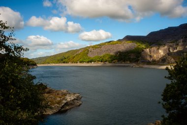 Snowdonia, Galler 2025.09.20 Kayalık tepelerle çevrili sakin dağ gölü ve yumuşak bulutlu açık mavi gökyüzünün altındaki orman yamaçları. 