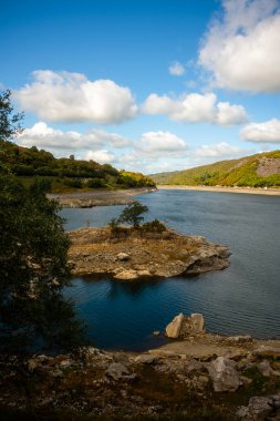 Kardan Donya, Galler 2025.09.20 Sakin mavi sularla çevrili küçük kayalık ada ve dağılmış bulutlarla kaplı parlak gökyüzünün altındaki uzak ormanlık tepeler.  