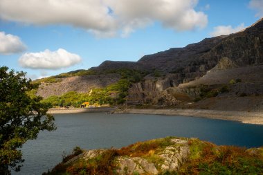 Kardan Donya, Galler 2025.09.20 Huzurlu mavi göl kayalık yamaçlarla çevrili ve yeşil gökyüzü altında dağınık beyaz bulutlar. 