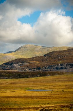 Snowdonia, Galler 2025.09.21 Sunlit altın vadisi ve kayalık dağ sırtı kalın kayan bulutlar ve mavi gökyüzü parçalarının altında sonbahar ışığı altında. 