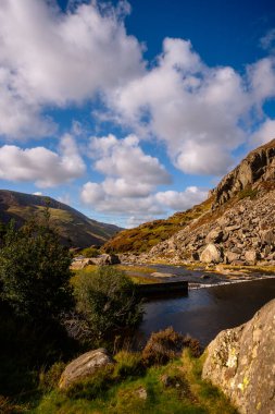 Snowdonia, Galler 2025.09.21 Nehir, sonbahar tepeleri ve açık mavi gökyüzü üzerindeki parlak bulutlarla çevrili güneşli kayalık bir vadide esiyor.. 