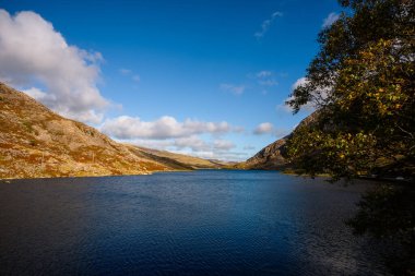 Kardan Donya, Galler 2025.09.21 Barış dolu dağ gölü sonbahar renkli tepeler ve kayalık yamaçlar altında dağınık mavi bulutlarla çevrili.. 