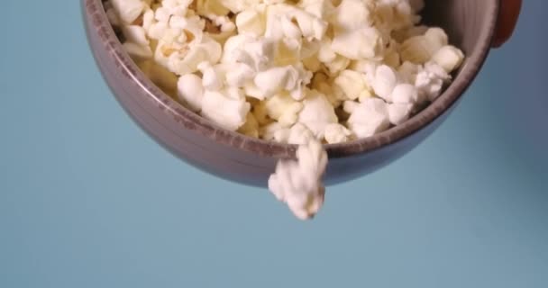 Popcorn falling from bowl on blue background, perfect snack for watching movie at home. — Stock Video