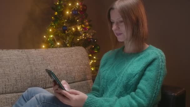 Жінка розмовляє по мобільному телефону в різдвяний вечір з ялинкою на фоні. Пандемія святкування на новий рік з мобільним телефоном без гостей . — стокове відео