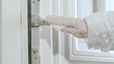 Koruyucu eldivenli bir kadın kapı koluna dezenfektan sürüyor ve özel bir peçeteyle siliyor.. 